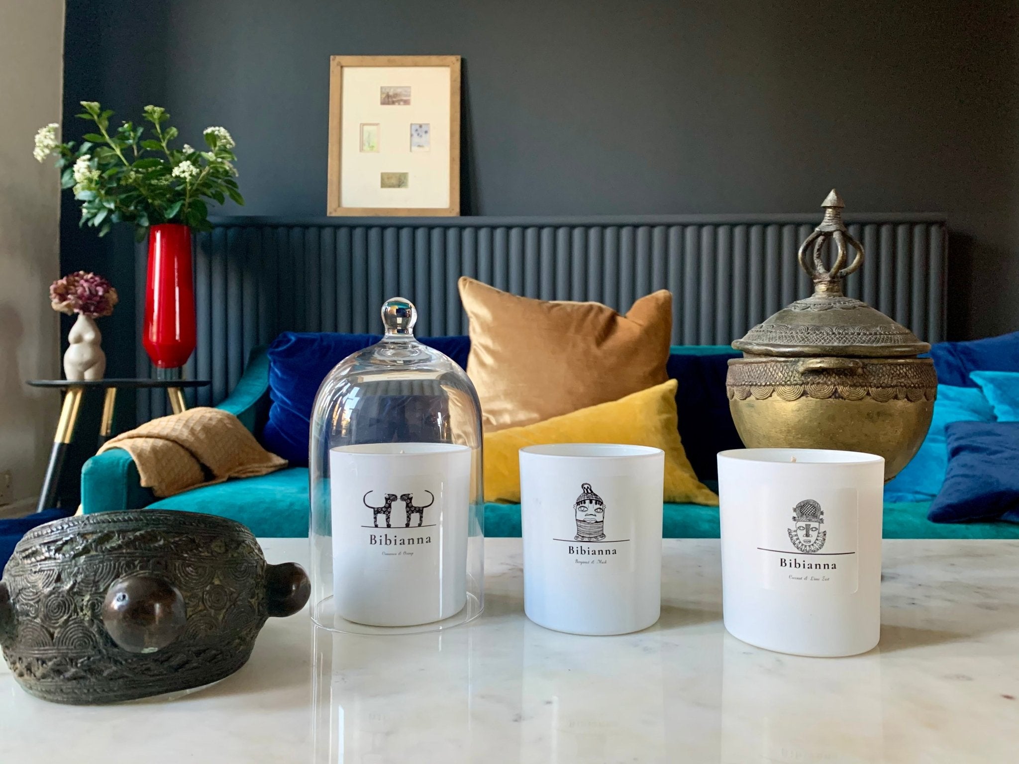 Three white candles labeled “Bibiana” sit on a marble table with decorative items, including a glass cloche, bronze turtle, and ornate container. In the background are colorful pillows on a blue sofa and a red vase with flowers.