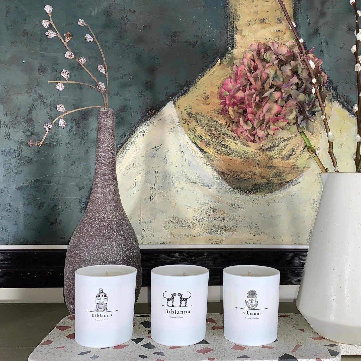 Three white candles labeled Bibiana are placed on a speckled surface in front of a painting featuring a floral bouquet and a woman’s torso. Two tall vases, one with buds and one with dried hydrangeas, stand on either side.