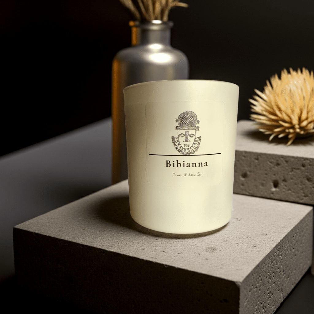 The Bibianna Scented Candle - Coconut & Lime by bibianna.co.uk features a frosted glass holder with a face design, displayed on a gray stone slab with a silver vase and straw decor for an elegant, luxurious touch.
