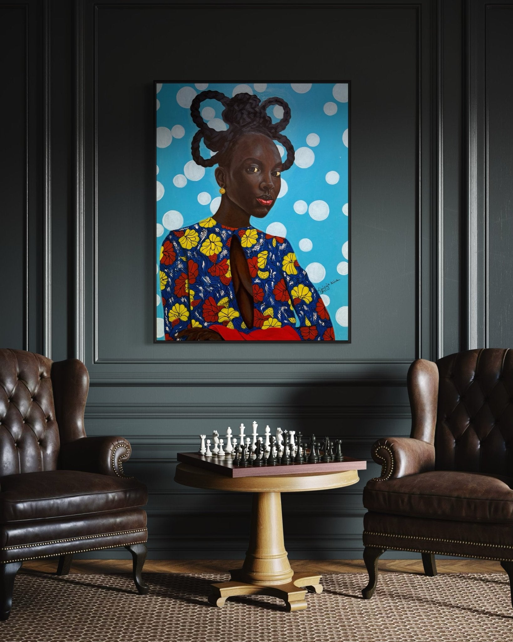 Two brown leather armchairs face a round table with a chess set in a dark-paneled room. Above hangs "Enigmatic Beauty: 'Lady'" by Ajenifuja Abiodun, reflecting contemporary African art's influence in this sophisticated setting.