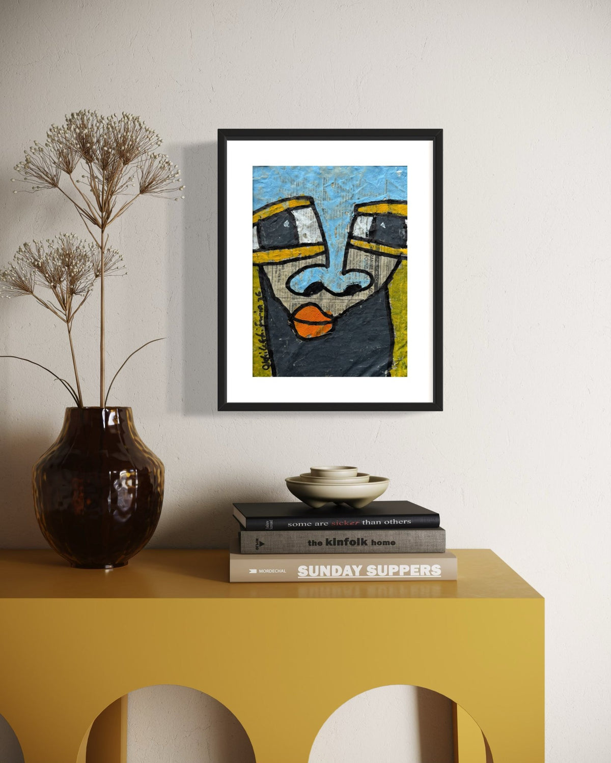 A framed abstract art print featuring a stylized face with blue, yellow, black, and orange details, displayed on a light beige wall above a yellow console table with a dark brown vase and books.
