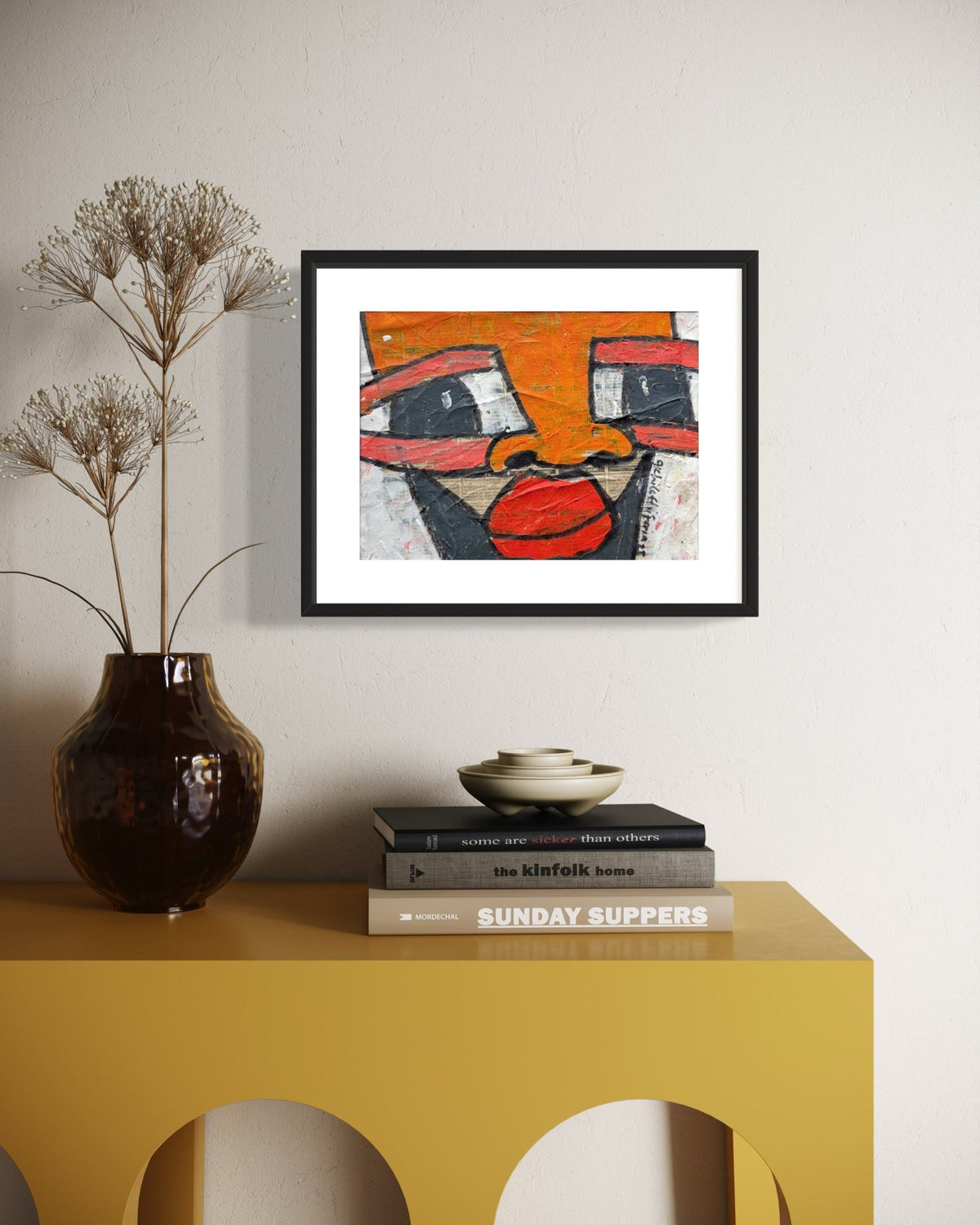 A framed abstract art print featuring a stylized face in orange, red, black, and white, displayed on a light beige wall above a yellow console table with books and a vase.