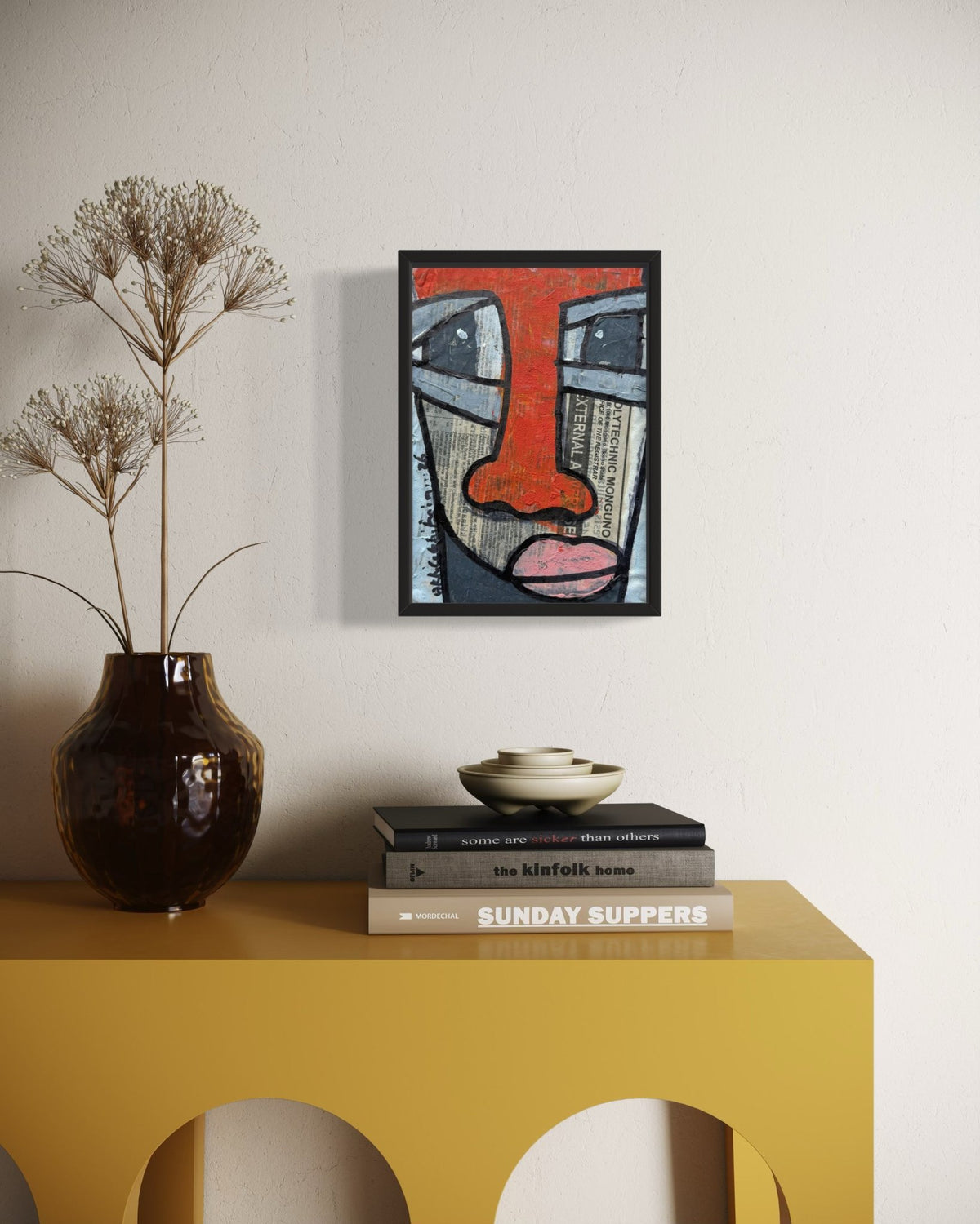 A framed abstract art print featuring a stylized face in red, gray, black, and pink tones, displayed on a white wall above a yellow console table with a dark brown vase and books.