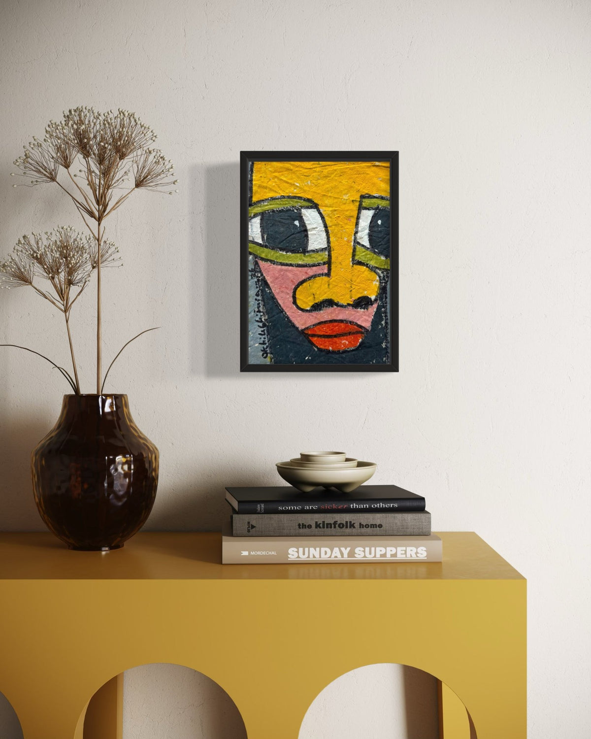 A framed abstract art print featuring a stylized face in yellow, green, pink, and red tones, displayed on a white wall above a mustard yellow console table with a dark brown vase and books.