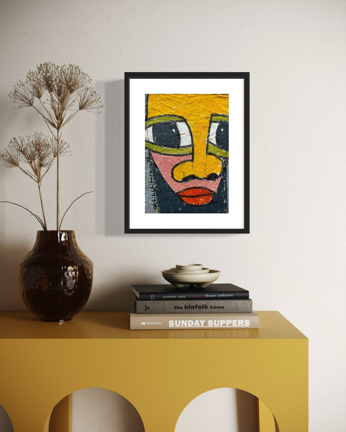 A framed art print featuring a colorful, textured face illustration hangs on a light beige wall above a yellow console table. The print is styled in a modern interior setting alongside a dark brown ceramic vase with dried grass and a stack of books.