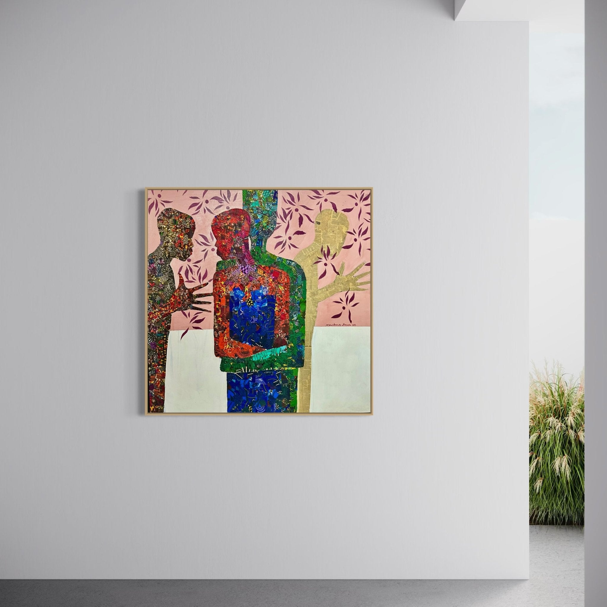 “Self” by Seye Morakinyo, a colorful mixed media on canvas artwork, shows three human-like figures and a bird with red, green, and blue patterns and pink floral shapes in the background. It hangs on a white wall with a plant visible outside.