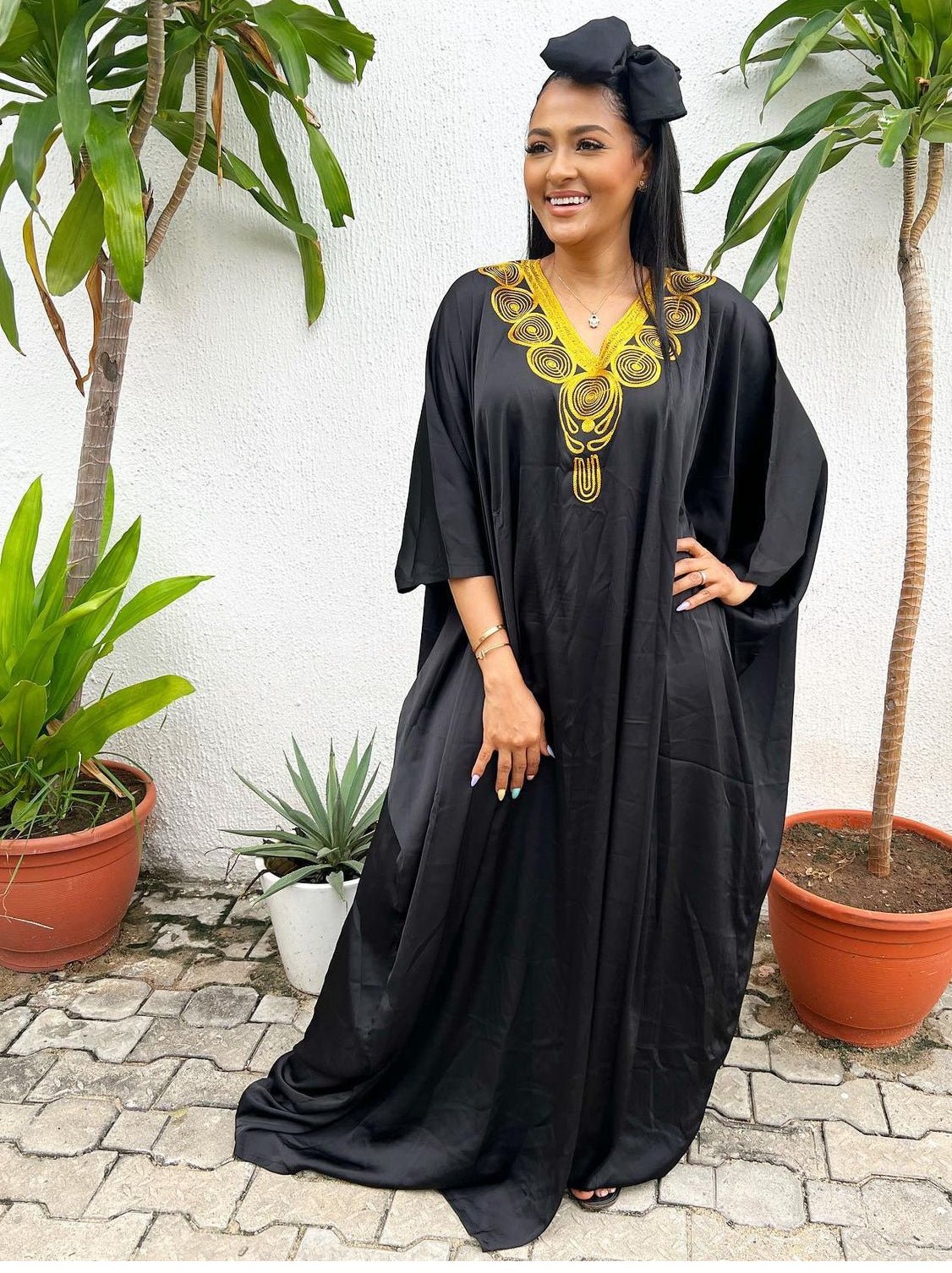 A woman stands outdoors on a stone path, smiling in the Silk Embroidered Bubu - Yellow by bibianna.co.uk with a matching headwrap. Green potted plants flank her against a white wall, showcasing this elegant African resort wear.