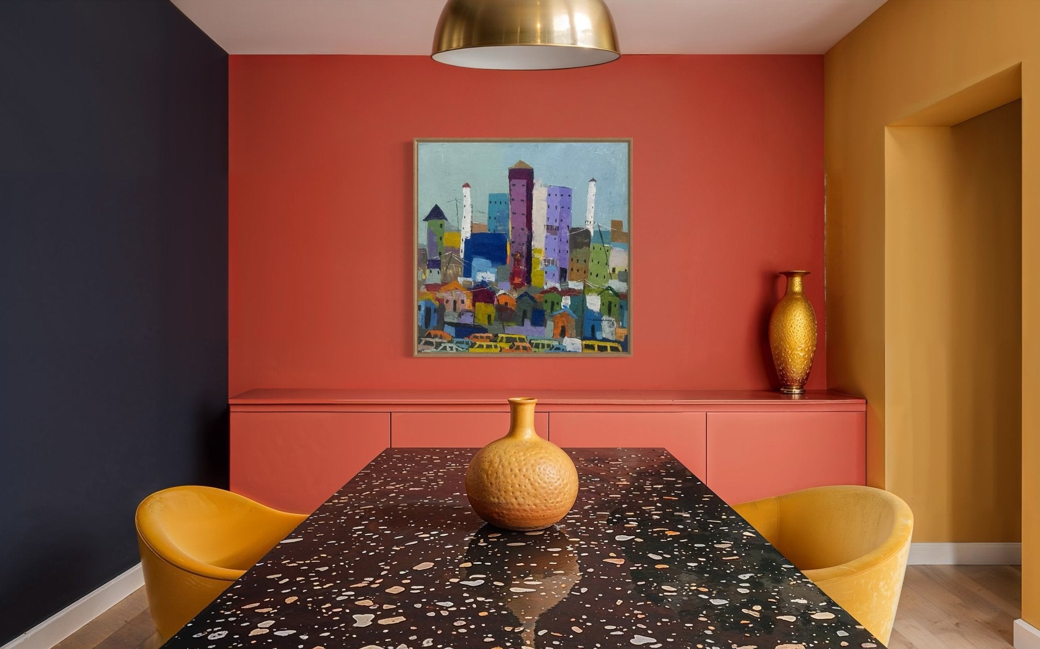A modern dining room with a terrazzo table, yellow chairs, and a gold vase. Navy, coral, and mustard yellow walls showcase "Somewhere In Lagos City" by Achike Anayo above the table under a striking gold pendant light.