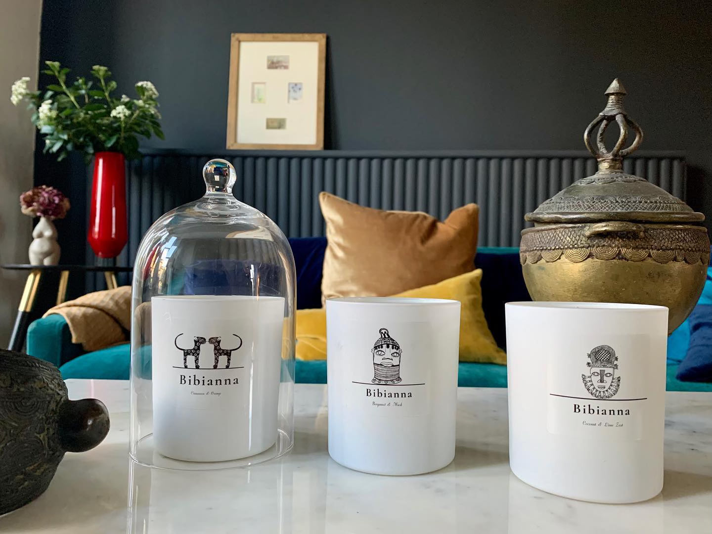 Three white Bibian­na candles with black illustrations on a marble table; one candle is under a glass cloche. Behind them are colorful cushions, a red vase with flowers, and a decorative bronze container.