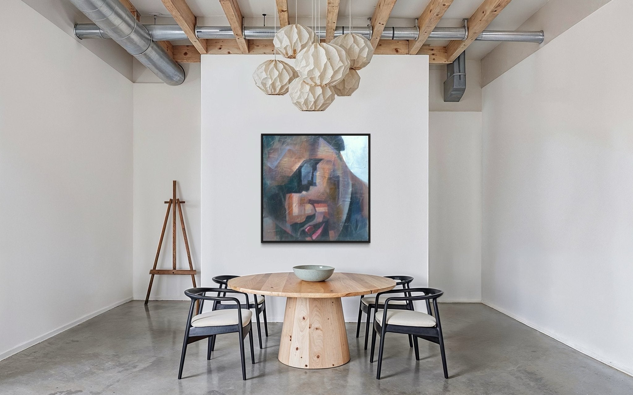 A modern dining area with a round wooden table, four black chairs, "Peace" Textured Painting by Christopher Ankeli on a white wall, a bowl on the table, an easel in the corner, and pendant lights from a wooden beam ceiling.