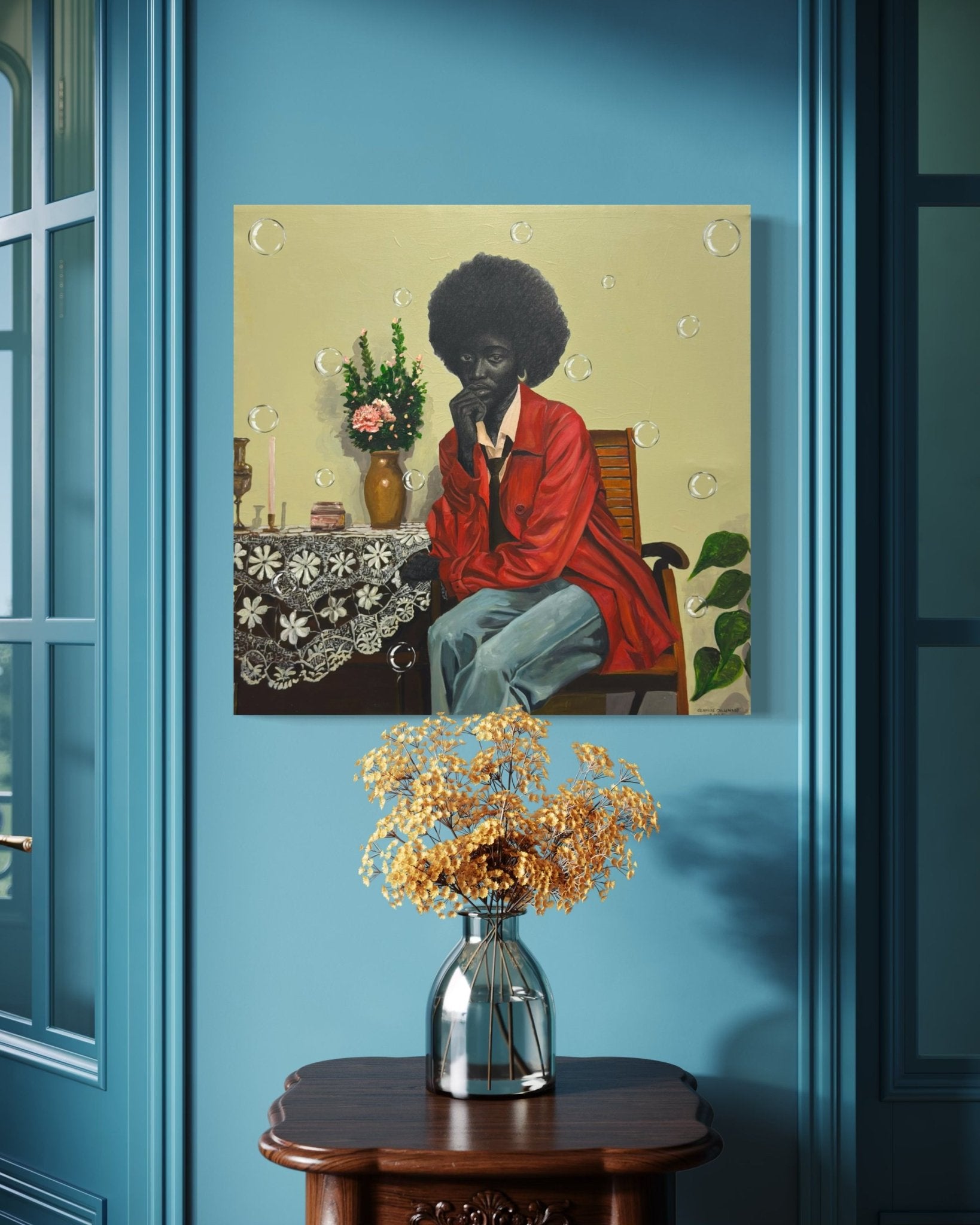 “The Presence Of Silence” by Olamide Ogunade, inspired by contemporary African art, depicts a figure with an afro and a red shirt among flowers. It hangs on a blue wall above a small wooden table with a vase of dried flowers.