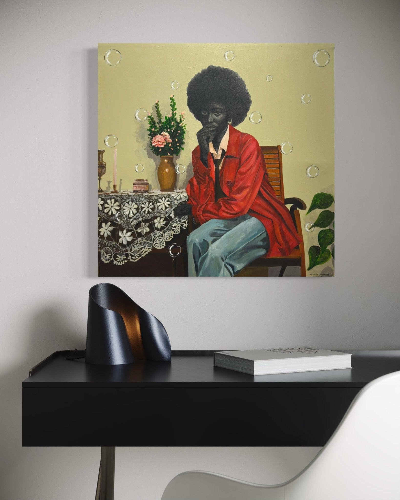 The Presence Of Silence" by Olamide Ogunade—a framed work inspired by contemporary African art, depicting a figure in a red jacket with flowers and bubbles—hangs above a modern black desk with sculptural decor, a book, and a white chair.