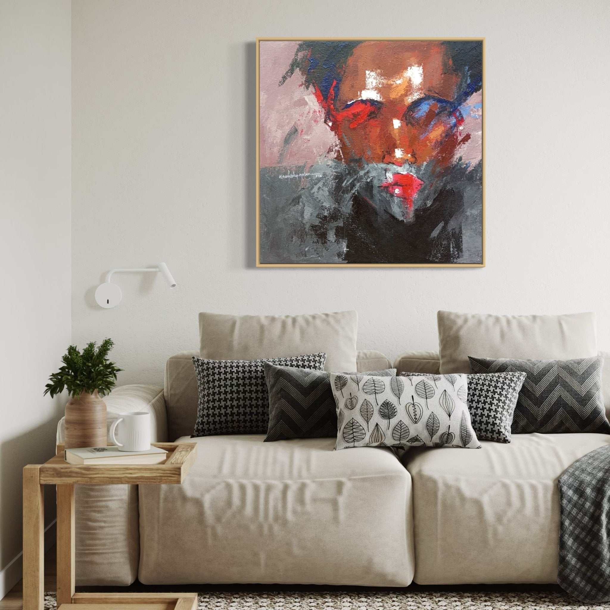 A living room with a beige sofa, patterned black and white cushions, a wooden side table with a plant and cup, and "Where I Am" (93 x 93cm) by Olasunkanmi Oyelusi hanging above the sofa.
